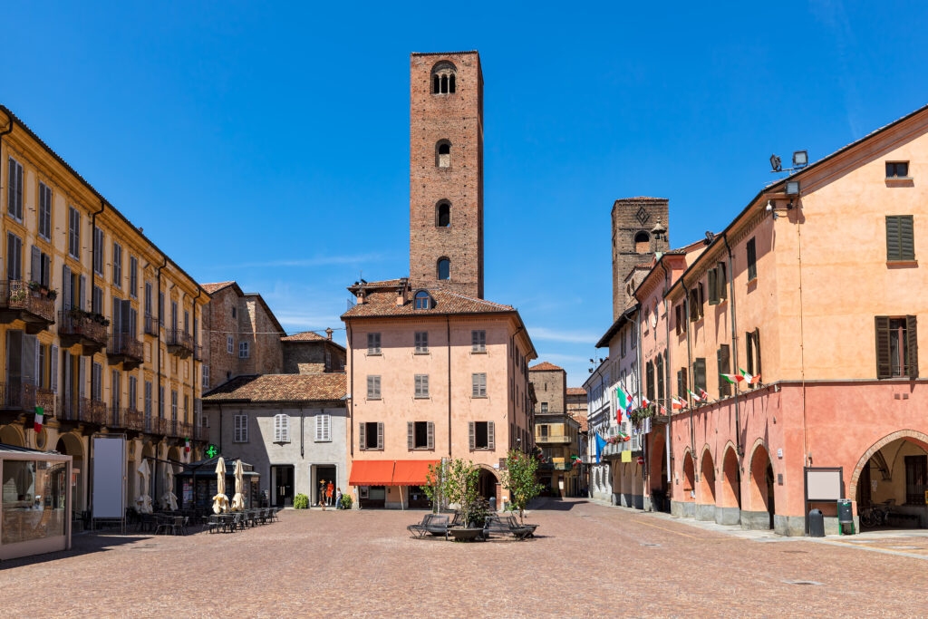 Cobblestone town square surrounded by typical colorful houses and medieval towers in Alba, Piedmont, Northern Italy