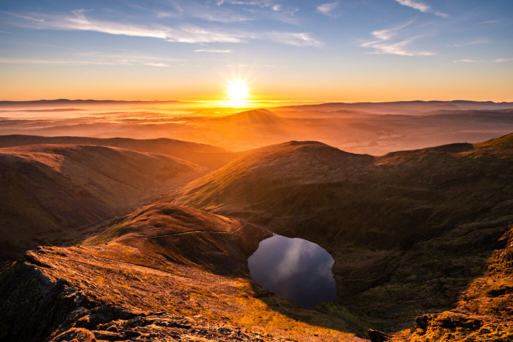 Stunning orange yellow sunrise from Sharp Edge on Blencathra, Lake District, Cumbria in Spring Summer with reflection in lake tarn and cloud inversion over mountains in the distance