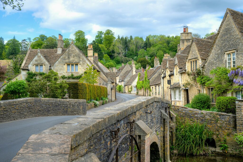 Charming view of traditional stone cottages and a bridge in Castle Combe, England, surrounded by lush greenery under a partly cloudy sky.