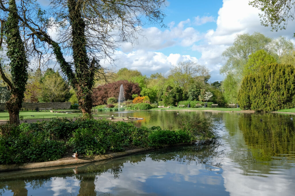 Pinner Memorial Park, Pinner, Middlesex, UK. Photo taken on a sunny (partially cloudy) spring day, showing lake with fountain, trees and green foliage.