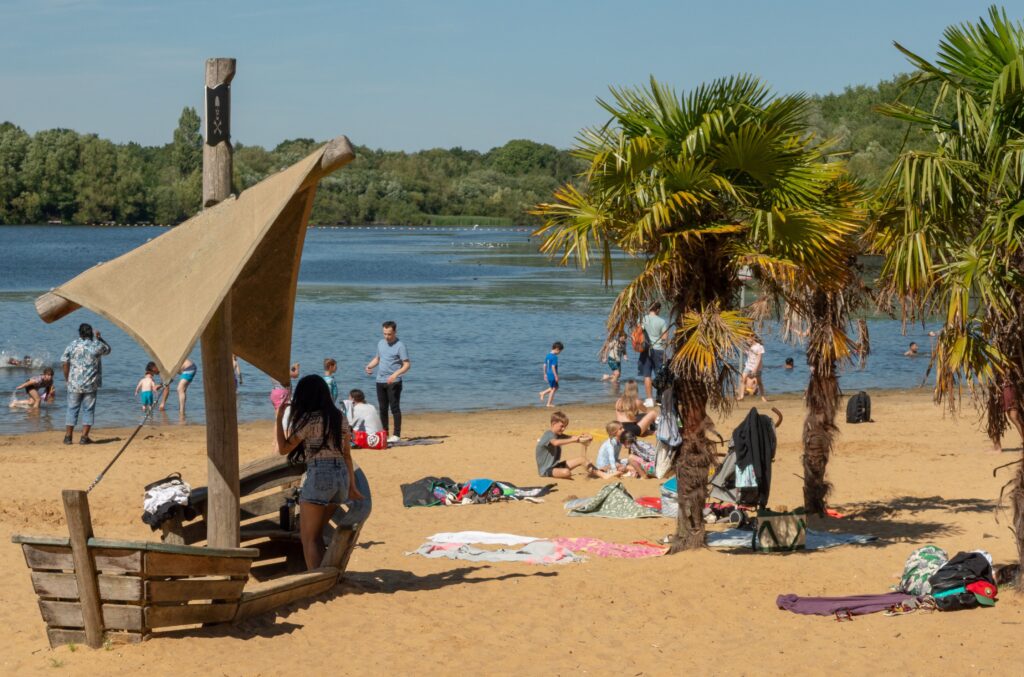 Tranquil sandy beach at Ruislip Lido with palm trees, a wooden pirate ship, and a scenic lake. People enjoying the peaceful lakeside setting