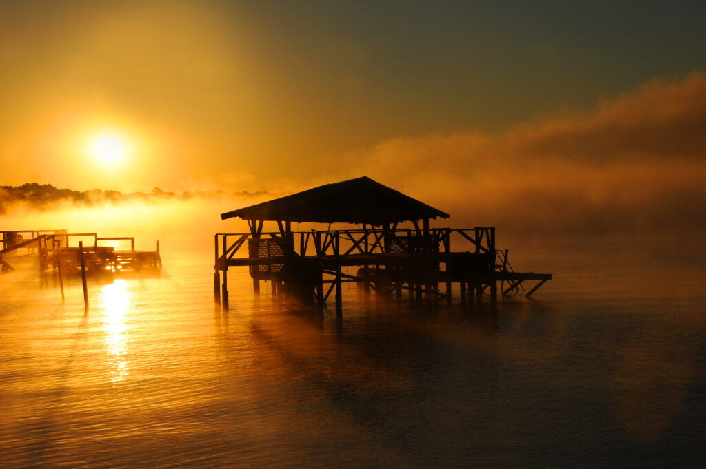 Early morning mist rises from Lake Chicot in Lake Village, Arkansas. Wooden dock and boat house are silhouetted. Golden light covers lake