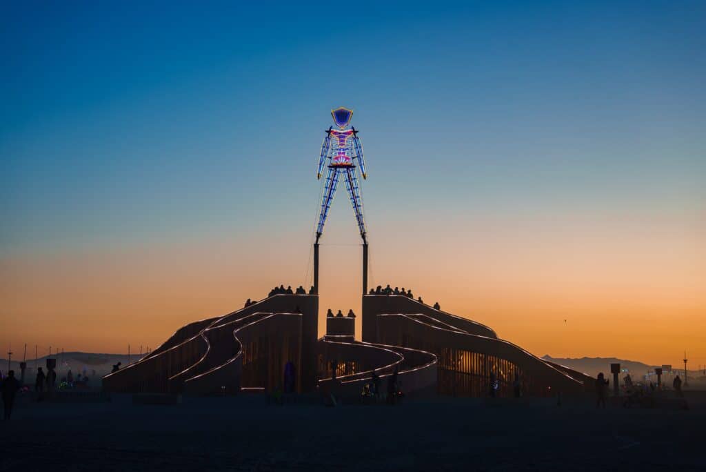 Nevada, USA. A large, colorful man sculpture stands atop a wooden structure in the Black Rock Desert at sunset, surrounded by festival participants. Burning Man festival.