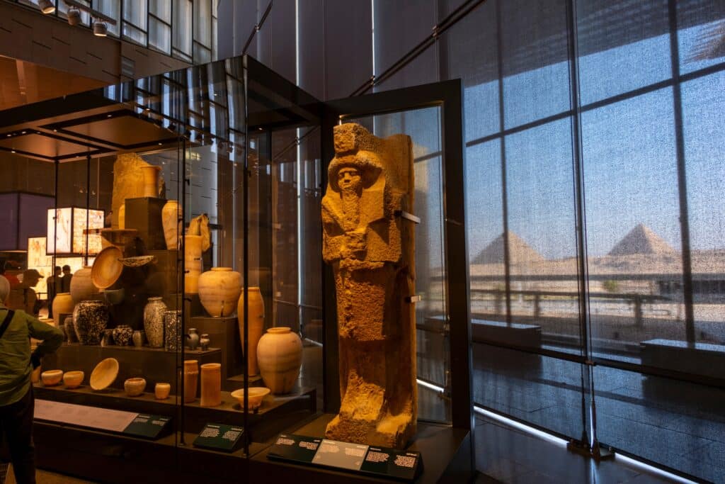 Giza, Egypt: Ancient Egyptian artifacts on display inside the Grand Egyptian Museum (GEM), with the Giza Pyramids visible through the windows.