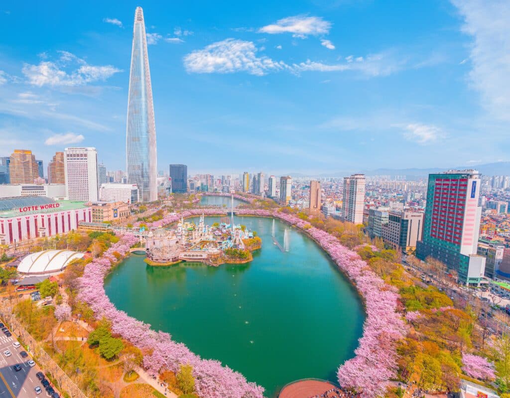 Lotte World Amusement Park in Seoul, South Korea, surrounded by full-bloom cherry blossoms and the Lotte World Tower. A clear sky enhances the beauty of spring