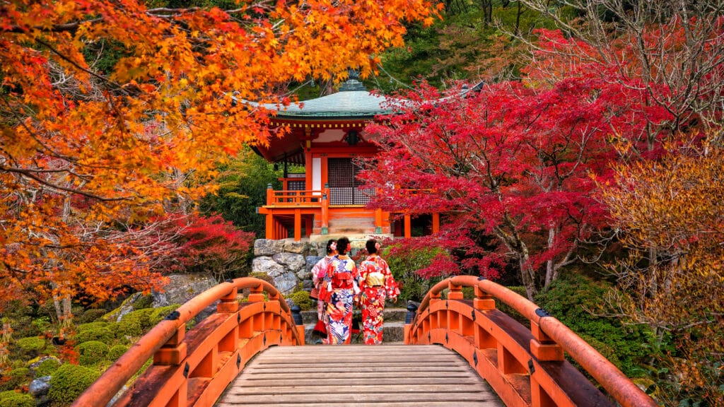 Visitors in kimono crossing a red bridge surrounded by autumn leaves at a temple garden in Japan