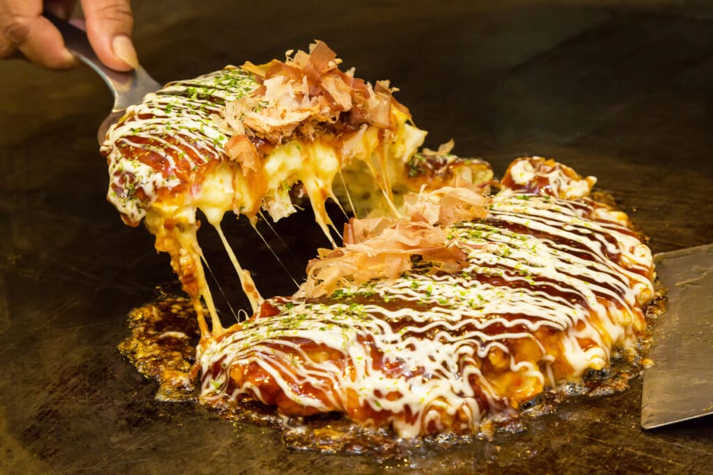 Okonomiyaki topped with sauce, mayonnaise and bonito flakes on a grill