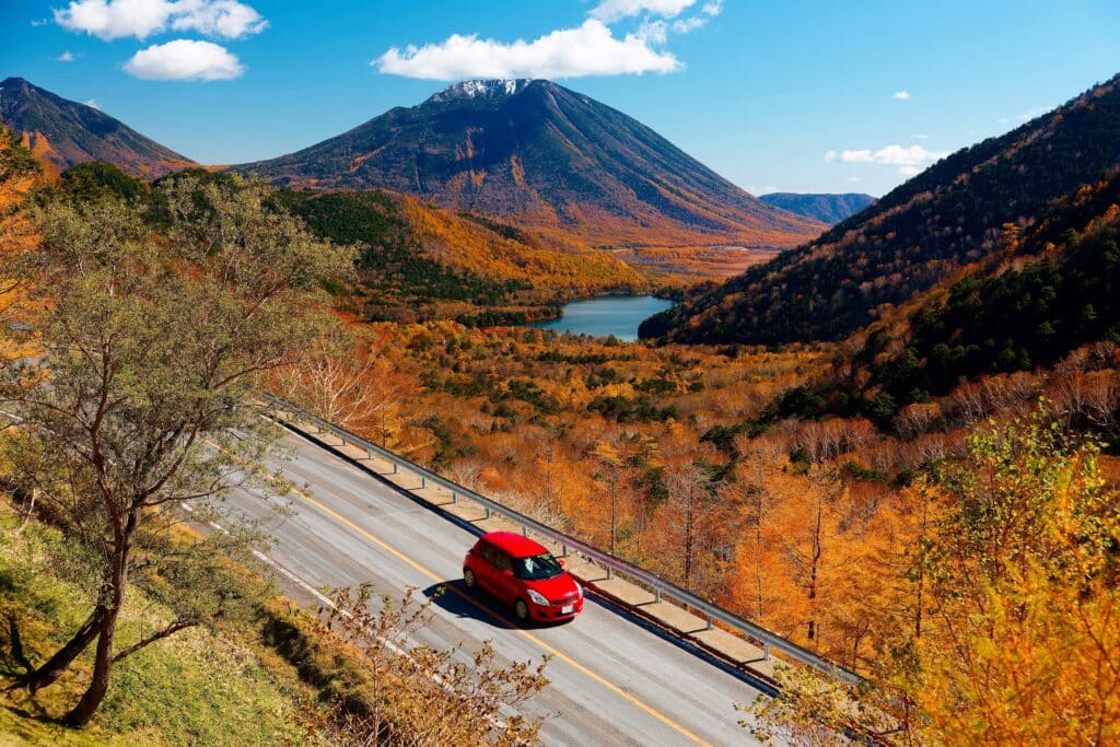 Red rental car driving through a scenic mountain road in rural Japan during autumn