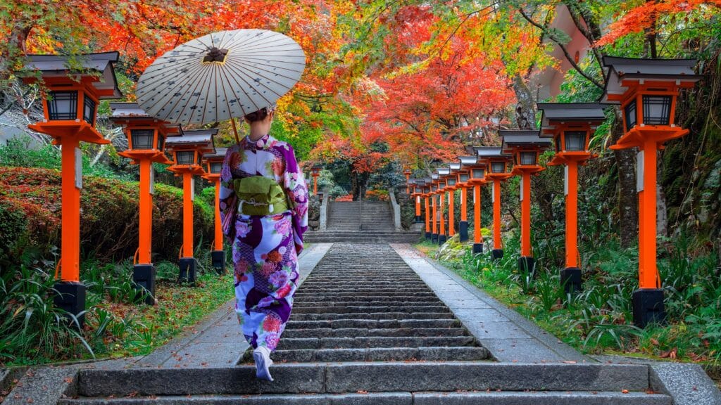 Visitor in traditional kimono walking along a lantern-lined temple path surrounded by colorful autumn foliage in Kyoto