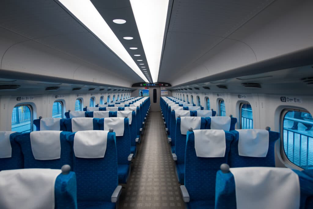 Interior of a Japanese Shinkansen train with rows of blue seats, white headrest covers, and a central aisle