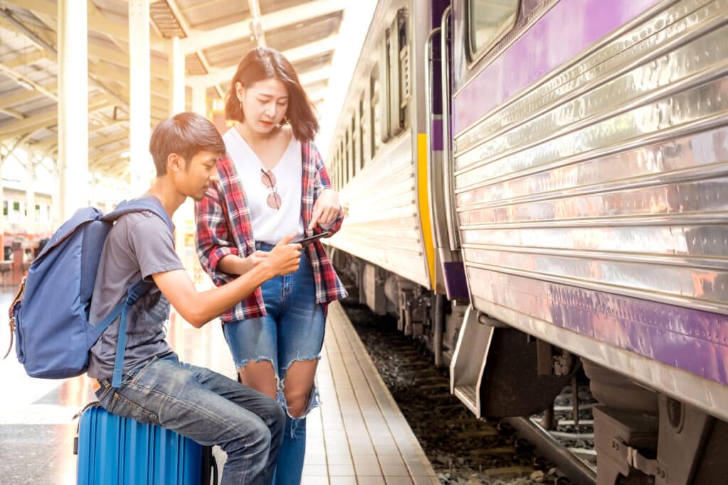 Two travelers checking their phone beside a train while carrying luggage at a railway station