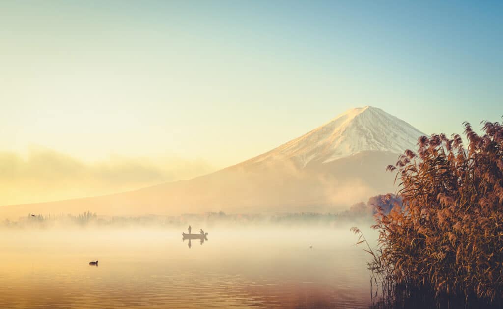 Mount Fuji rising above a mist-covered lake in warm morning light with a small boat on the water