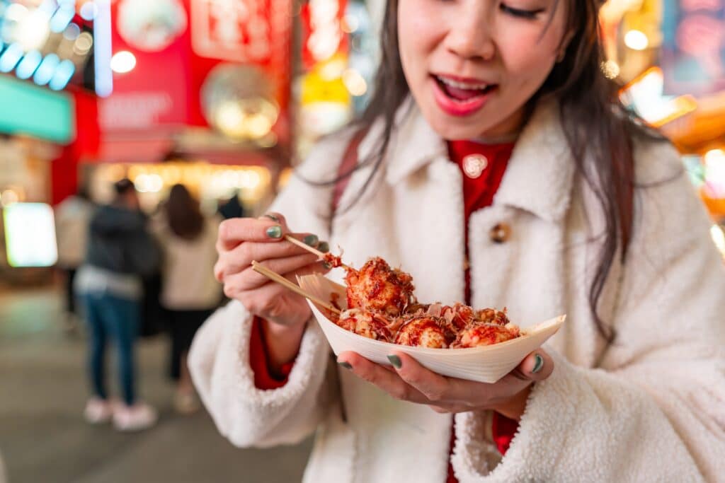 Woman eating freshly prepared takoyaki from a paper tray in a colorful Japanese street-food district